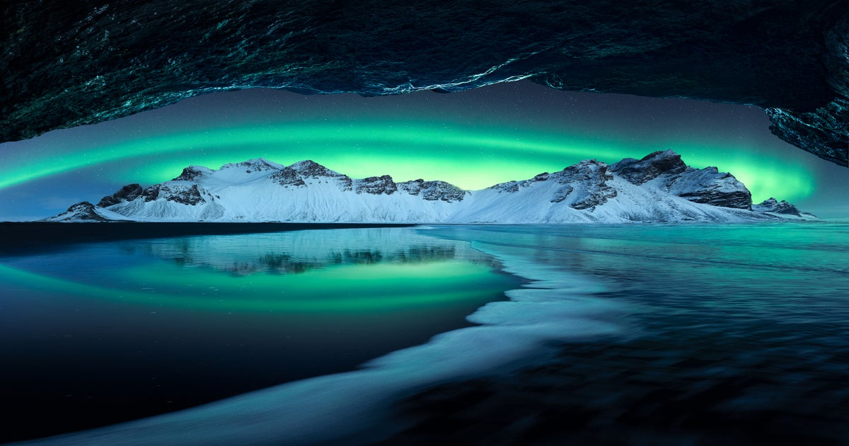 Green aurora over a snowy mountain ridge reflected on a wet black sand beach